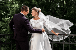Braut und Bräutigam stehen auf einem Balkon, umgeben von üppigem Grün, und verkörpern die perfekte Vorstellung einer Hochzeit. Die Braut trägt ein weißes Spitzenkleid mit fließendem Schleier, während der Bräutigam einen dunklen Anzug trägt. Sie blicken sich liebevoll an, während der Schleier sanft im Wind weht.