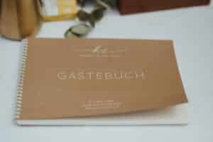 Ein spiralgebundenes Gästebuch mit braunem Einband und dem Titel „Gästebuch“ erinnert an den besonderen Anlass der Hochzeit von Kimberly und Christian am 4. Juni 2022. Es ist perfekt für ihre Hochzeitsfeier und ruht elegant auf einer weißen Tischdecke mit sanftem Dekor im Hintergrund.