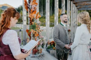 Ein Paar steht während einer rustikalen Hochzeitszeremonie im Freien. Als Teil ihrer Hochzeitsplanung trägt der Bräutigam ein traditionelles Outfit und die Braut eine weiße Jacke. Eine Frau amtiert mit einem Buch in der Hand. Holzbalken und leuchtende Herbstblumen schmücken die Kulisse.