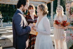 Bei einer wunderschön geplanten Hochzeit tauschen Braut und Bräutigam in einer Zeremonie im Freien die Ringe aus. Die Braut lächelt freudig in ihrem weißen Kleid, während der Bräutigam im Anzug neben ihr steht. Brautjungfern mit Blumen stehen in der Nähe, während eine Frau in einem gemusterten Kleid diesen freudigen Moment der Hochzeit zelebriert.