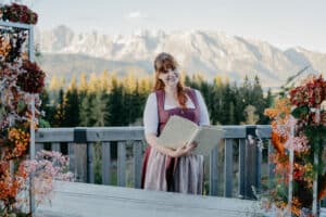 Eine Frau in traditioneller Kleidung steht auf einer Holzterrasse und hält ein offenes Buch in der Hand. Vielleicht träumt sie von ihrer bevorstehenden Hochzeit. Sie ist von bunten Blumen umgeben und hat einen malerischen Blick auf Bäume und Berge unter einem klaren Himmel, der die perfekte Kulisse für Hochzeiten bildet.