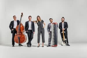 Eine Gruppe von fünf Musikern steht in einer Reihe vor einem schlichten Hintergrund und gibt damit den perfekten Ton für jede Hochzeit vor. Einer hält einen Kontrabass, ein anderer ein Keyboard, ein dritter eine Gitarre und die zentrale Figur stützt sich auf einen anderen Musiker. Sie sind in formelle Kleidung gekleidet, ideal für eine elegante Hochzeitsplanung.