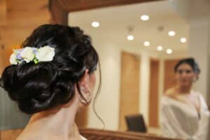 Eine Frau mit einer eleganten Hochsteckfrisur und Blumenschmuck betrachtet ihr Spiegelbild. Sie trägt ein weißes Gewand, perfekt für eine Hochzeit, und Ohrringe in einem sanft beleuchteten Raum mit Holzhintergrund.