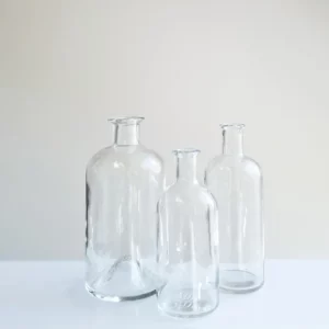 Drei klare Glasflaschen unterschiedlicher Höhe stehen auf einer weißen Fläche vor einem schlichten, hellen Hintergrund. Die leeren Flaschen sind in einer dreieckigen Formation angeordnet und eignen sich perfekt, um jeder Hochzeit einen Hauch von Eleganz zu verleihen oder als Teil Ihrer exquisiten Hochzeitsplanungsdekoration.