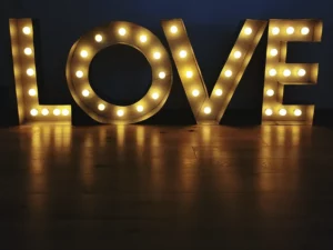 Große beleuchtete Buchstaben bilden „LOVE“ vor einem dunklen Hintergrund und fangen die Essenz der Hochzeitsmagie ein. Die leuchtenden Buchstaben werfen warme Reflexionen auf den darunterliegenden Holzboden und schaffen eine perfekte Kulisse für Heiratsträume.
