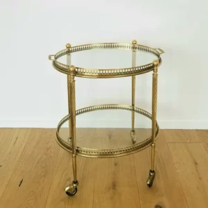 Ein zweistöckiger Barwagen aus Goldmetall im Vintage-Stil mit Glasplatte und Ablage, perfekt für elegante Hochzeitsfeiern. Er verfügt über aufwendige Details an den Kanten und zylindrische Beine mit kleinen Lenkrollen und steht anmutig auf einem Holzboden vor einer schlichten weißen Wand.