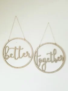 Zwei runde Wandbehänge aus Holz mit kursivem Text, perfekt für die Hochzeitsplanung. Auf dem linken Kreis steht „better“ und auf dem rechten „together“, elegant an dünnen Schnüren vor einem schlichten weißen Hintergrund aufgehängt. Ideale Dekoration, um Ihre Reise zur Hochzeit zu feiern.