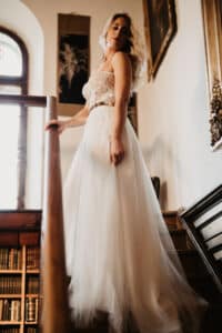 Eine Frau in einem fließenden weißen Kleid steht anmutig auf einer Treppe und blickt nach unten. Sie verkörpert die ruhige Eleganz einer Hochzeit. Sonnenlicht fällt durch ein nahes Fenster und hebt ihr blondes Haar hervor. Die Kulisse scheint eine elegante, antike Bibliothek mit Büchern in Regalen zu sein.