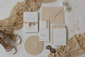 Eine flache Lage eleganter Hochzeitspapeterie in neutralen Tönen, darunter Einladungen, Umschläge, ein Menü, eine Tischnummer und Namenskarten. Beiger Stoff, mit Juwelen besetzte High Heels, Goldschmuck und cremefarbene Blütenblätter betonen das Arrangement.