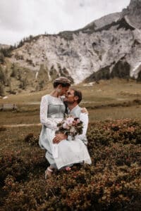 Ein Paar in traditioneller Kleidung sitzt liebevoll auf einer üppigen Bergwiese und verkörpert die Essenz einer perfekten Hochzeit. Die Frau hält einen Blumenstrauß vor felsigen Hügeln und verstreutem Grün unter einem bewölkten Himmel und fängt den Geist einer glückseligen Hochzeit ein.