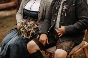 Ein Paar in traditioneller bayerischer Kleidung, möglicherweise mitten in der Hochzeitsplanung, sitzt nebeneinander. Die Frau trägt ein Dirndl und hält einen Strauß getrockneter Blumen, während der Mann Lederhosen und eine Strickjacke trägt. Sie sitzen im Freien auf Holzstühlen.