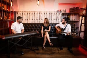 In einem gemütlichen Raum, der sich perfekt für die Hochzeitsplanung eignet, sorgen drei Musiker für die richtige Atmosphäre: ein Mann am Keyboard, eine Frau in der Mitte und ein weiterer Mann an der Gitarre. Sie sitzen auf einer dunklen Lederbank, umgeben von warmem Licht und dekorativen Holzpaneelen im Hintergrund.
