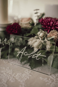 Ein durchsichtiges Acrylschild mit der kursiven Aufschrift „Acht“ steht auf einem Tisch und eignet sich perfekt als Hochzeitsdekoration. Dahinter verleiht ein Blumenarrangement mit roten und weißen Blumen, grünen Blättern und einer Spitzentischdecke jeder Hochzeitsfeier Eleganz.