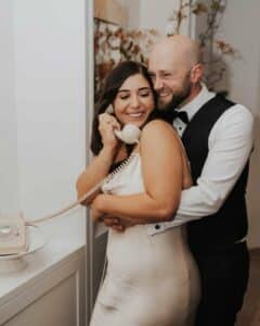 Ein lächelndes Paar umarmt sich, während die Frau ein Vintage-Telefon mit Wählscheibe in der Hand hält, das die Freude der Hochzeit verkörpert. In formeller Kleidung stehen sie vor einer hellen, mit zartem Laub geschmückten Wand und halten einen Moment fest, der sich anfühlt, als gehöre er zu ihrer perfekten Hochzeitsplanung.
