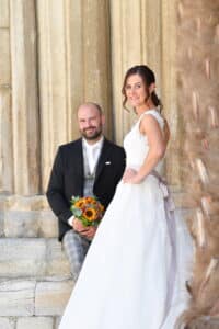 Eine Braut in einem weißen Kleid lächelt in die Kamera, während sie vor hohen Steinsäulen steht. Es ist ein perfekter Moment ihrer Hochzeit, als der Bräutigam, stilvoll gekleidet in einem dunklen Anzug und karierten Hosen, neben ihr sitzt und einen farbenfrohen Strauß aus Sonnenblumen und orangefarbenen Blumen hält.