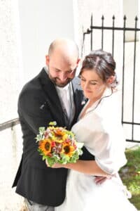 Braut und Bräutigam umarmen sich zärtlich im Freien neben einem schwarzen Metalltor und verkörpern damit die Essenz ihrer Hochzeit. Sie hält einen Strauß Sonnenblumen und kleine rote Blumen in der Hand und beide sind elegant gekleidet – der Bräutigam in einem schwarzen Anzug und die Braut in ihrem atemberaubenden weißen Kleid.