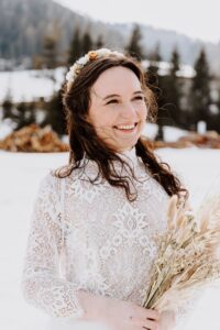 Eine lächelnde Frau in einem Spitzenkleid hält einen Strauß getrockneter Blumen in der Hand und verkörpert damit die Essenz des Heiratens. Sie steht in einer verschneiten Landschaft mit Bäumen im Hintergrund und strahlt Freude aus, während ihr Haar, geschmückt mit einem Blumenstirnband, sanft ihr Gesicht umrahmt – ein perfekter Moment auf dem Weg der Hochzeitsplanung.