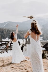 Zwei Frauen in weißen Spitzenkleidern stehen auf einem grasbedeckten Hügel, umgeben von schneebedeckten Bergen und Gebäuden. In einer freudigen Feier ihrer Hochzeit wirft eine Frau einen Blumenstrauß in die Luft, während die andere ihn auffangen will. So fängt sie den Geist des Heiratens in einem unvergesslichen Moment ein.