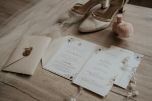 Ein Paar elfenbeinfarbene Brautschuhe, ein Hochzeitsschleier und ein versiegelter Brief liegen auf einer Holzoberfläche. Daneben befinden sich elegante Hochzeitseinladungen mit Blumenmotiven und eine Flasche rosa Nagellack mit einem flauschigen Applikator – perfekte Details für Ihre Hochzeitsvorbereitungen.