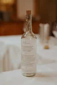 Eine Glasflasche mit Korken, perfekt für die Hochzeitsplanung, wird als Menühalter auf einer weißen Tischdecke verwendet. Das Menü ist auf Papier in der Flasche gedruckt. Im Hintergrund schaffen verschwommenes Geschirr und sanfte Beleuchtung eine warme Atmosphäre, ideal für eine intime Hochzeit.