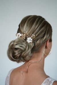 Eine Frau mit blondem Haar, das zu einem eleganten, tiefen Dutt gestylt ist, geschmückt mit dekorativen goldenen Blättern und weißen Blumen – ideal für eine Hochzeit. Sie trägt große Creolen und ein Spitzentop. Der Hintergrund ist in einer schlichten hellen Farbe gehalten, perfekt, um Erinnerungen an die Hochzeit festzuhalten.