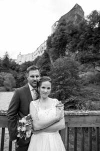 Ein Schwarzweißfoto zeigt ein Paar mitten in der Hochzeitsplanung auf einer Holzbrücke. Die Frau im weißen Kleid hält einen Blumenstrauß, während der Mann im Anzug hinter ihr steht. Im Hintergrund sind ein großes Gebäude und Bäume auf einem Hügel zu sehen, was ihren perfekten Hochzeitsplanungsmoment noch verstärkt.