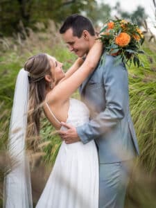 Braut und Bräutigam umarmen sich freudig auf einer Wiese und verkörpern damit die Essenz der Hochzeit. Die Braut in ihrem weißen Kleid und Schleier hält einen Strauß orangefarbener Blumen in der Hand. Der Bräutigam, in einen grauen Anzug gekleidet, lächelt zärtlich, während sie einander in die Augen blicken.