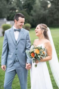 Braut und Bräutigam lachen und halten sich während ihrer Hochzeit auf einer Wiese an den Händen. Der Bräutigam trägt einen hellgrauen Anzug mit Fliege, während die Braut in einem weißen Kleid glänzt und einen Strauß orangefarbener und grüner Blumen in den Händen hält.