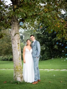 Ein Paar steht an seinem Hochzeitstag unter einem Baum in einem üppigen, grünen Park. Die Braut trägt ein weißes Kleid und der Bräutigam einen hellgrauen Anzug. Sie lächeln und sind von kleinen schwebenden Seifenblasen umgeben.