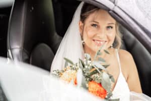 Eine fröhliche Braut mit Schleier sitzt in einem Auto und hält einen Strauß mit orangefarbenen Blumen und Grünpflanzen in der Hand, was die Freude am Heiraten verkörpert. Sie lächelt in die Kamera und schafft so eine warme, fröhliche Atmosphäre. Das Fenster ist leicht unscharf, was ihren strahlenden Gesichtsausdruck noch mehr in den Mittelpunkt rückt.