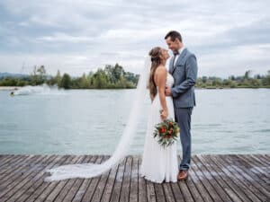 Braut und Bräutigam stehen auf einem Holzsteg an einem ruhigen See, umarmen sich und schauen einander in die Augen. Die Braut trägt ein weißes Kleid und hält einen Blumenstrauß, während der Bräutigam einen grauen Anzug trägt. Als Teil ihrer Hochzeit verleiht der bedeckte Himmel mit den Bäumen im Hintergrund Romantik.