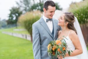 Braut und Bräutigam lächeln sich bei ihrer Hochzeit im Freien an. Die Braut hält einen Strauß orangefarbener Blumen in der Hand und trägt ein weißes Kleid und einen Schleier. Der Bräutigam trägt einen hellgrauen Anzug mit Fliege. Im Hintergrund sind Grünflächen und ein Weg zu sehen, die perfekte Hochzeitsplanung verkörpern.
