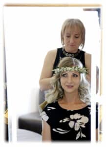 Eine Frau mit lockigem blondem Haar und einem Blumenkranz sitzt lächelnd vor einem Spiegel, während eine Friseurin in einem schwarzen ärmellosen Oberteil ihre Frisur perfektioniert. Im Salon herrscht geschäftiges Treiben bei der Hochzeitsplanung.