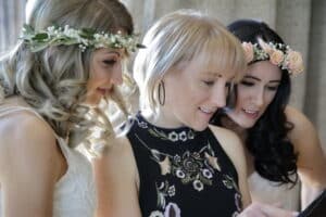 Drei Frauen stehen dicht beieinander, lächeln und schauen auf ein Smartphone. Zwei von ihnen tragen Blumenstirnbänder und weiße Outfits, was darauf hindeutet, dass sie mitten in der Hochzeitsplanung stecken, während die dritte ein schwarzes Kleid mit Blumenmuster und Creolen trägt. Sie scheinen einen angenehmen Moment zusammen zu genießen.