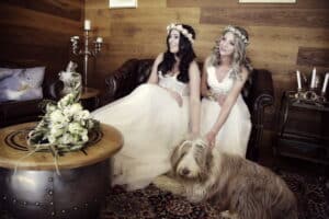 Zwei Frauen in weißen Kleidern und Blumenkränzen, die vielleicht ihre Hochzeit planen, sitzen auf einem Ledersofa, lächeln und streicheln zärtlich einen zotteligen Hund auf dem Boden. Ein Strauß weißer Blumen schmückt den runden Holztisch vor ihnen, der vor einer rustikalen, holzgetäfelten Wand steht.