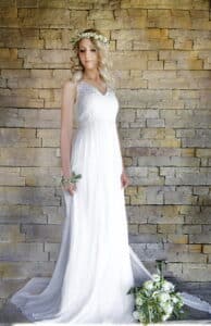 Eine Braut in einem weißen Spitzenkleid steht vor einer Steinmauer und verkörpert die Essenz einer Märchenhochzeit. Sie trägt eine Blumenkrone und hält einen Strauß weißer Blumen in der Hand. Ihr blondes Haar ist in lockeren Wellen gestylt und ihre Augen sind sanft geschlossen, was den heiteren Moment des Heiratsglücks einfängt.