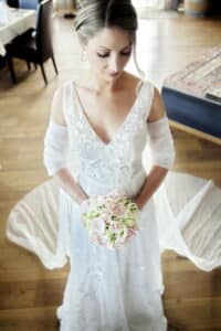 Eine Braut in einem weißen Spitzenkleid hält einen Strauß rosa und weißer Rosen in der Hand und verkörpert damit die Essenz einer Hochzeit. Sie steht auf einem Holzboden, hinter ihr hängt ein durchsichtiger Schleier. Mit elegant gestyltem Haar blickt sie in den sanft beleuchteten Raum hinunter und fängt die heitere Schönheit des Heiratens ein.