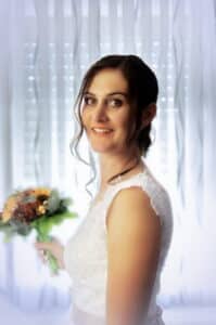 Eine Braut in einem weißen Spitzenkleid hält einen Blumenstrauß in der Hand und strahlt in die Kamera. Vor transparenten Vorhängen, durch die das Licht fällt, ist ein perfekter Moment der Freude festgehalten, der während ihrer sorgfältig geplanten Hochzeit entstand.