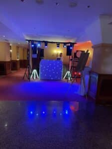 In einem Hotelflur steht ein DJ-Pult mit bunten Lichtern, perfekt für eine Hochzeitsfeier. Eine Person steht hinter dem Pult, während helle blaue Lichter den Boden beleuchten und die festliche Atmosphäre verstärken. Dekorative Beleuchtung und Ausstattung umgeben den Bereich, der sowohl mit Teppichboden als auch mit Fliesen ausgelegt ist.