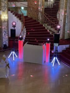 Vor einer großen Treppe mit rotem Teppich ist ein DJ-Pult mit leuchtenden Lichtern für eine Hochzeit aufgebaut. Zwei große Lautsprecher und Beleuchtungsgeräte flankieren das Pult. Der Veranstaltungsort, perfekt für alle, die sich intensiv mit Hochzeitsplanung beschäftigen, besticht durch kunstvolles Dekor, darunter Kronleuchter und Marmorwände.