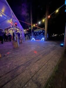 Eine Nachtszene mit bunten Lichterketten und einem beleuchteten DJ-Pult, umgeben von Bäumen, bildet die Bühne für eine zauberhafte Hochzeit. Ein Holzdeck führt zu einer Baldachinkonstruktion auf der linken Seite, wo sich die Leute zum Feiern versammeln. Der Bereich leuchtet in verschiedenen Farbtönen der dekorativen Lichter, perfekt für Hochzeiten.