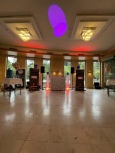 Ein geräumiger Raum, der für eine Veranstaltung eingerichtet ist, perfekt für eine Hochzeit, mit einem DJ-Pult mit leuchtenden LED-Lichtern und Lautsprechern. Auf den Tischen an den Seiten stehen verschiedene Dekorationen, darunter metallische Luftballons. Große Fenster bieten einen Blick auf das Grün draußen, während bunte Deckenleuchten für Atmosphäre sorgen.