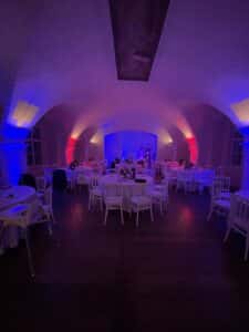 Ein schwach beleuchteter Bankettsaal mit gewölbten Decken, perfekt für eine Hochzeit, wird von blauen und roten Lichtern beleuchtet. Weiße runde Tische umgeben den Raum, während der zentrale Tisch, geschmückt mit Blumen und Kerzen, das elegante Ambiente verstärkt. Perfekt für diejenigen, die gerade mitten in der Hochzeitsplanung sind.