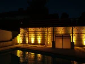 Ein schwach beleuchteter Außenpoolbereich bei Nacht, perfekt für Hochzeitsfeiern, mit warmen gelben Lichtern, die eine strukturierte Wand und gewölbte Paneele beleuchten. Schatten und Reflexionen schaffen eine ruhige Atmosphäre, ideal für Hochzeitsplanung unter den Sternen.