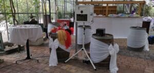 Eine rustikale Inneneinrichtung für eine Hochzeit mit in weißen Stoff eingewickelten Weinfässern, einer Fotokabine und einem Tisch mit bunten Perücken. Es gibt eine wunderschön dekorierte Kulisse mit Grünpflanzen und verschiedenen ausgestellten Foto-Requisiten, perfekt für Ihre Hochzeitsplanung.