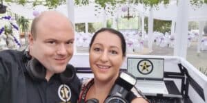 Ein Mann und eine Frau, beide mit Kopfhörern, lächeln in die Kamera. Die Frau hält eine Kamera mit Blitzlicht. Hinter ihnen ist ein dekorierter Saal mit weißen und violetten Stühlen und großen Fenstern, der die fröhliche Atmosphäre einer Hochzeitsfeier suggeriert.
