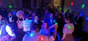 Es entfaltet sich eine lebhafte Tanzszene auf der Hochzeitsfläche, mit einer Gruppe gut gekleideter Gäste, die die Freude des Anlasses auskosten. Bunte Lichter erhellen den Raum und tragen zur festlichen Atmosphäre bei. Eine Braut in der Mitte lächelt und tanzt voller Energie und verkörpert einen Moment perfekter Hochzeitsplanung.