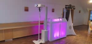 Ein tragbares Event-Setup, das sich perfekt für Hochzeitsfeiern eignet, besteht aus einem Projektor auf einem Ständer und einer transparenten LED-beleuchteten Kabine, die violett leuchtet. In der Nähe steht ein weißes dreieckiges Banner mit einem Logo auf dem Holzboden eines Innenraums, ideal für eine reibungslose Hochzeitsplanung.