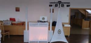 Ein für eine Hochzeit konzipiertes DJ-Setup umfasst Lautsprecher, einen Laptop und Beleuchtungsgeräte in einem Raum. Ein weißes Tuch mit einem Logo verleiht Eleganz, während ein Holztisch mit Körben den Hintergrund schmückt. Auf der linken Seite ist teilweise ein Schild sichtbar, das die festliche Atmosphäre verstärkt.