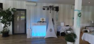 Ein dekorierter Veranstaltungsort, der sich perfekt für eine Hochzeit eignet, verfügt über eine DJ-Kabine mit Beleuchtungsanlage und Lautsprechern. Weiße und blaue Akzente, darunter herzförmige Ornamente, schmücken den Raum. Pflanzen und moderne Möbel vervollständigen das elegante Ambiente und machen es ideal für Ihren besonderen Tag der Hochzeit.